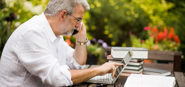 A person outdoors on their laptop and answering the phone with books scattered around