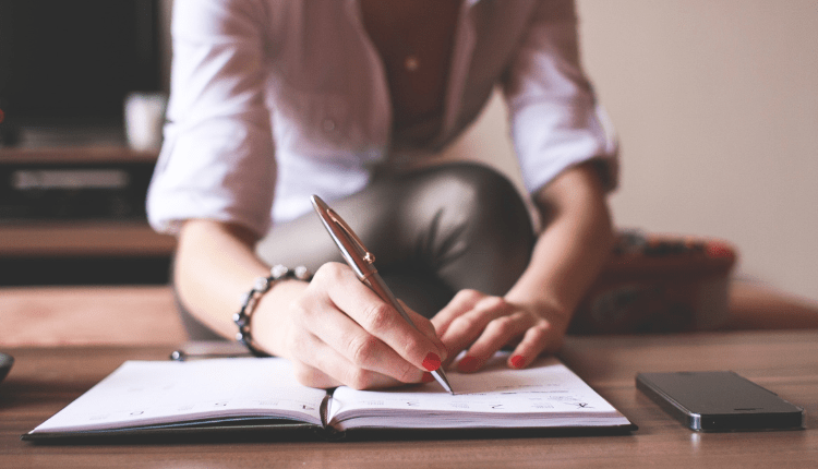 A person writing in a notebook at their desk with an iPhone to the side